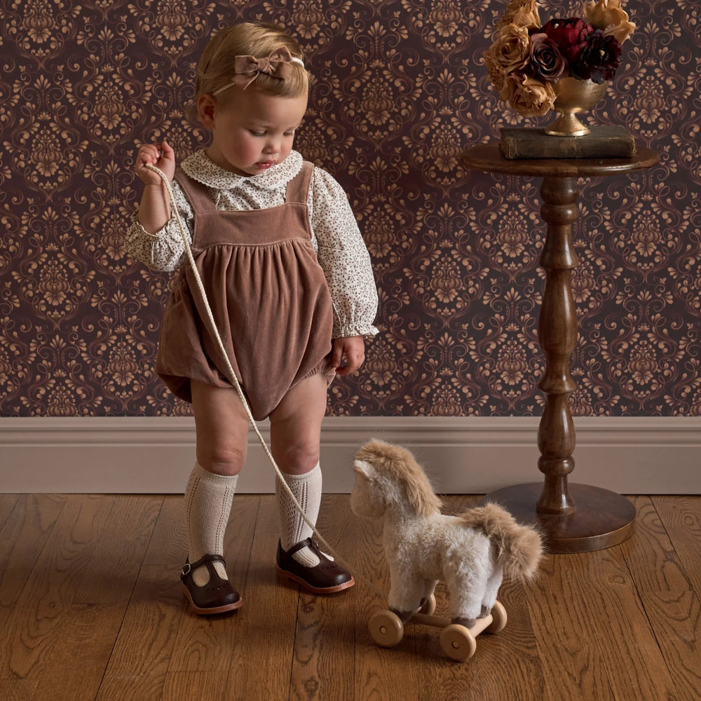 Child in a brown bubble standing next to a wooden horse toy in a room with patterned wallpaper.