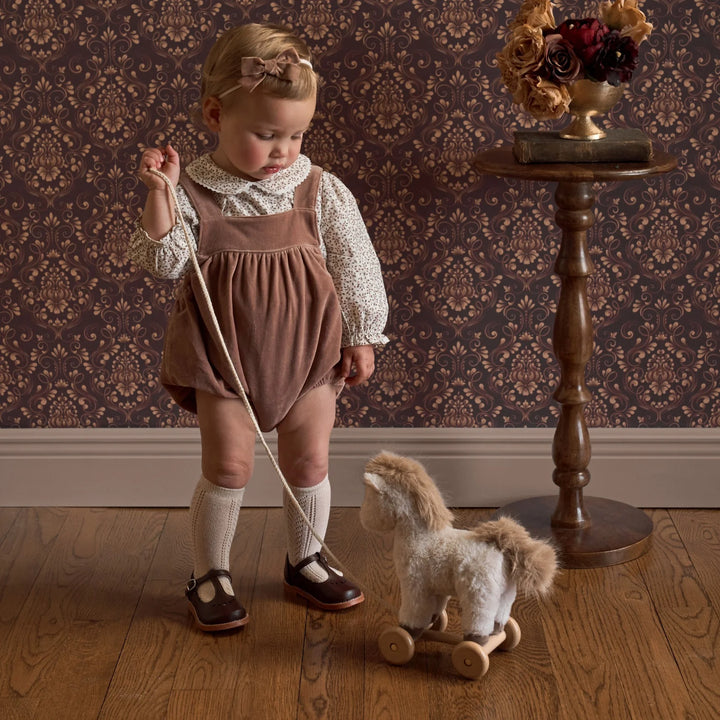 Child in a brown bubble standing next to a wooden horse toy in a room with patterned wallpaper.