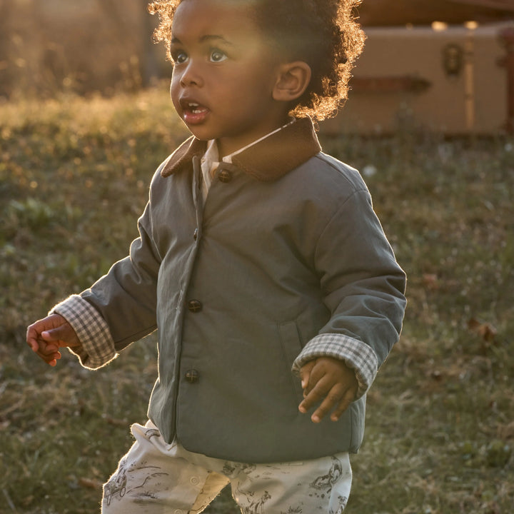 A toddler wearing a dark gray button-front jacket with a brown corduroy collar and white patterned pants, standing outdoors in warm golden-hour light.