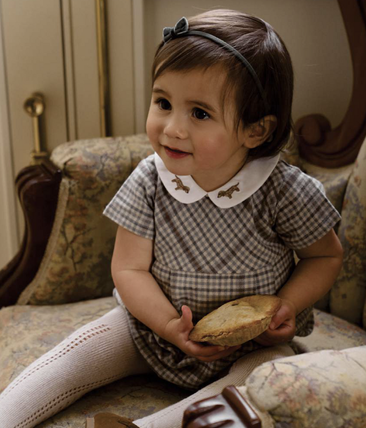 A toddler girl wearing a brown and white gingham dress with a white Peter Pan collar and white knit tights, sitting on a vintage floral armchair and holding a small pie.