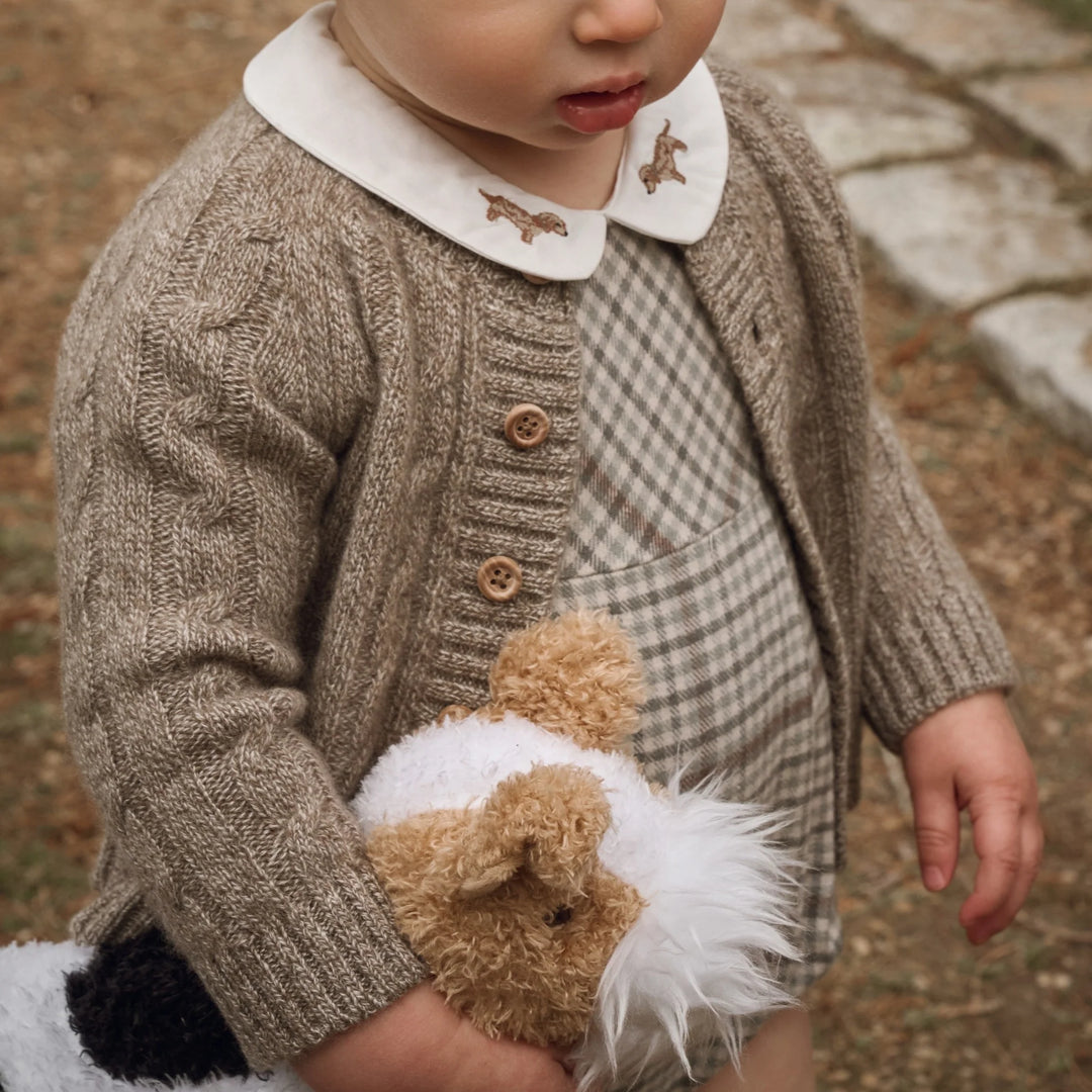 A close-up of a baby wearing a textured taupe knit cardigan with a white Peter Pan collar decorated with embroidered brown animals, over a beige and gray plaid romper. The baby is holding a plush toy in an outdoor setting.