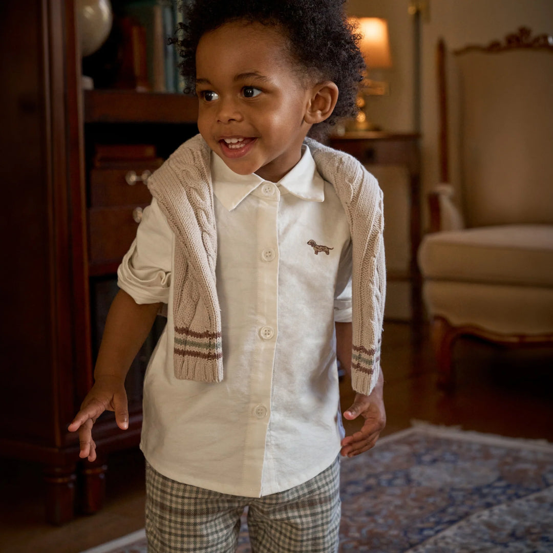 Child wearing a light-colored shirt with a small logo and plaid pants in a room with furniture.