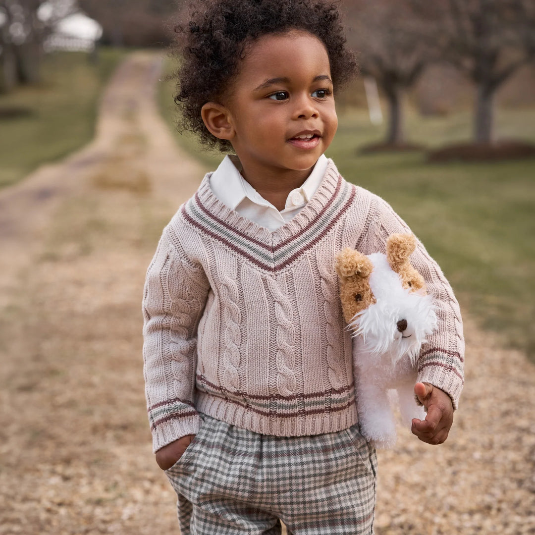 A young child standing outdoors on a gravel path wearing a light pink cable-knit sweater over a white collared shirt, with plaid pants and holding a small white and tan stuffed dog.
