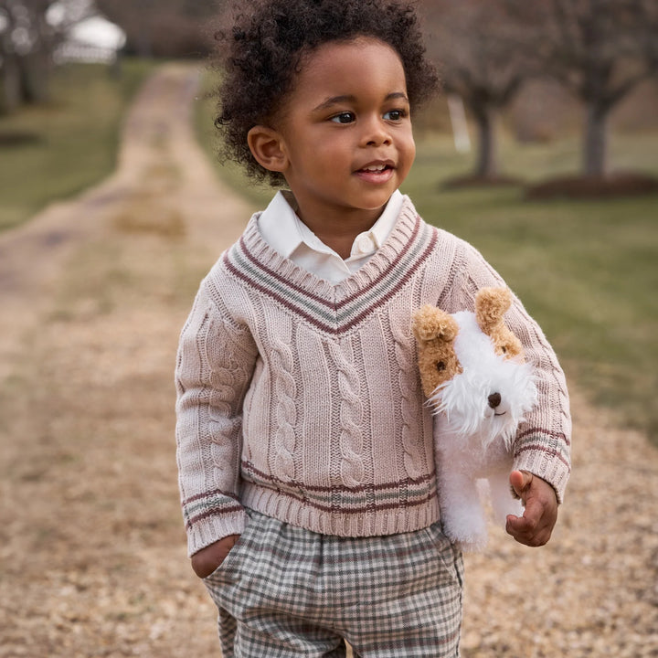 A young child standing outdoors on a gravel path wearing a light pink cable-knit sweater over a white collared shirt, with plaid pants and holding a small white and tan stuffed dog.