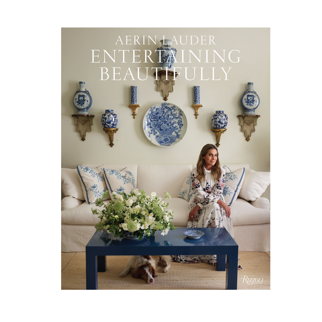 A book cover for 'Entertaining Beautifully' by Aerin Lauder, featuring a woman sitting on a beige sofa in a decorated living room with blue-and-white porcelain decor, a blue coffee table, and a dog underneath.