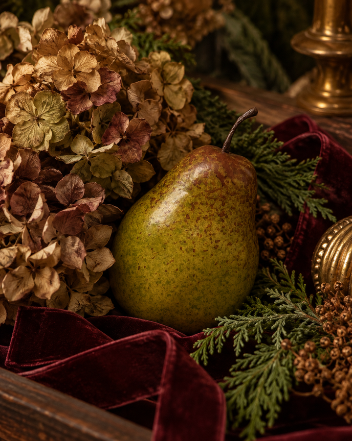 A green speckled pear nestled in a rustic arrangement with dried flowers, evergreen branches, a deep burgundy velvet cloth, and gold-toned decorative accents.