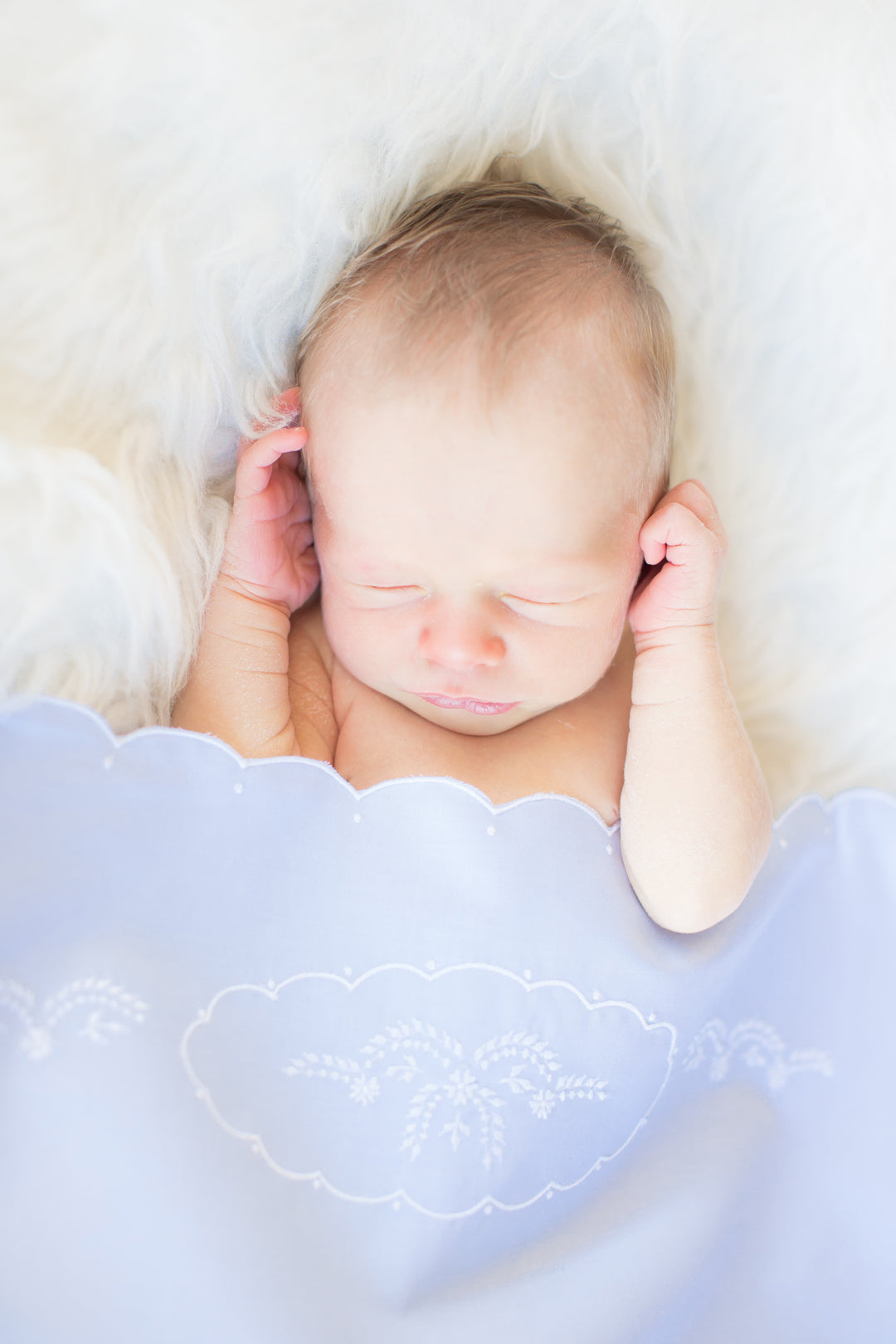 A sleeping newborn baby wrapped in a light blue blanket with white embroidered detailing, resting on a fluffy white faux fur surface.