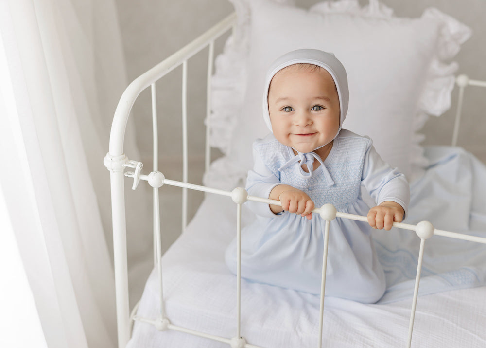 A baby wearing a light blue long-sleeve dress and matching bonnet sits on a white metal crib, holding the rails with both hands against a soft white background.