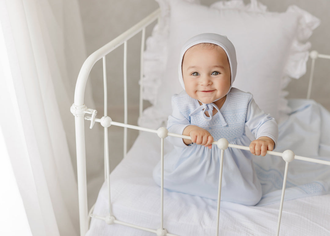 A baby wearing a light blue long-sleeve dress and matching bonnet sits on a white metal crib, holding the rails with both hands against a soft white background.