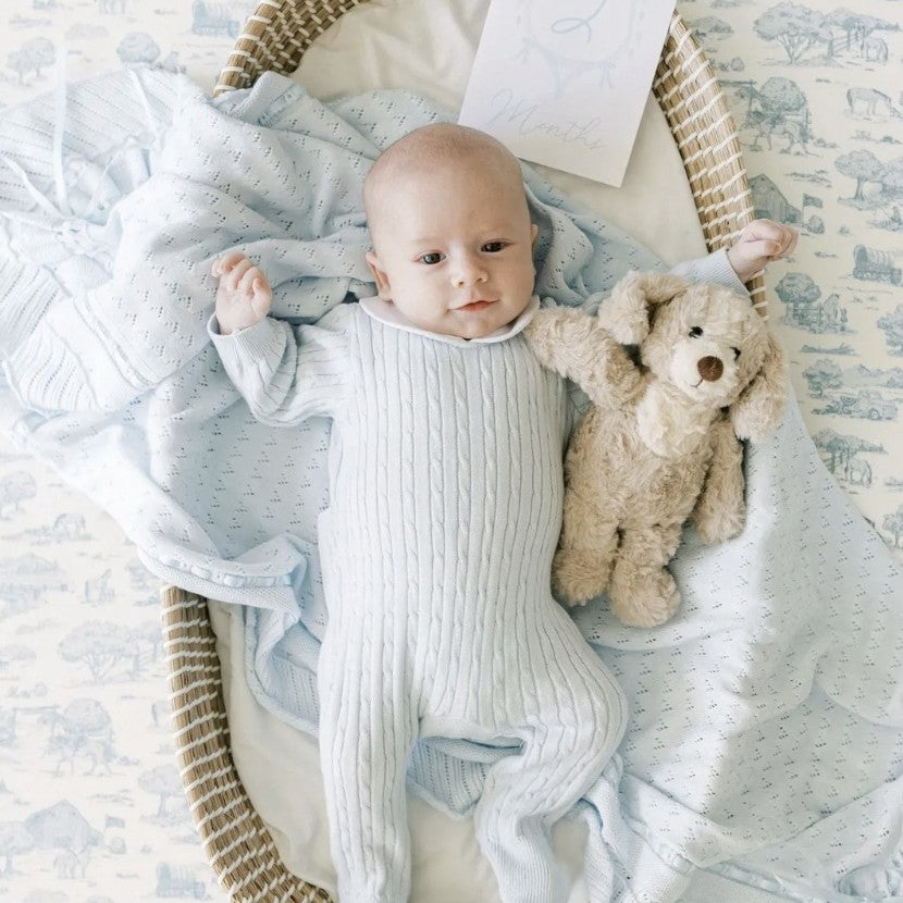 A baby lying in a woven bassinet, wrapped in a light blue ribbed footed sleeper and a pale blue patterned blanket, with a beige stuffed bunny toy beside them.