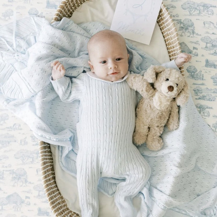 A baby lying in a woven bassinet, wrapped in a light blue ribbed footed sleeper and a pale blue patterned blanket, with a beige stuffed bunny toy beside them.