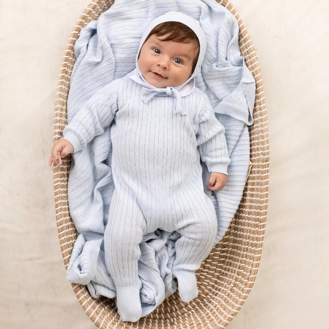 A baby wearing a light blue ribbed footed sleeper and bonnet, lying on a striped cream and beige woven mat with a matching blue blanket.