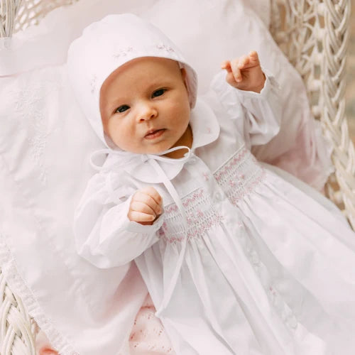 Baby in a white embroidered dress and bonnet sitting in a wicker chair.