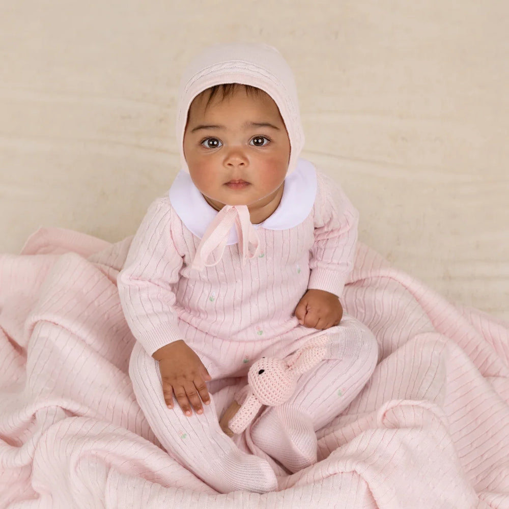 A baby wearing a light pink ribbed knit one-piece outfit with a white collar and matching bonnet, sitting on a soft pink blanket and holding a small pink stuffed bunny.