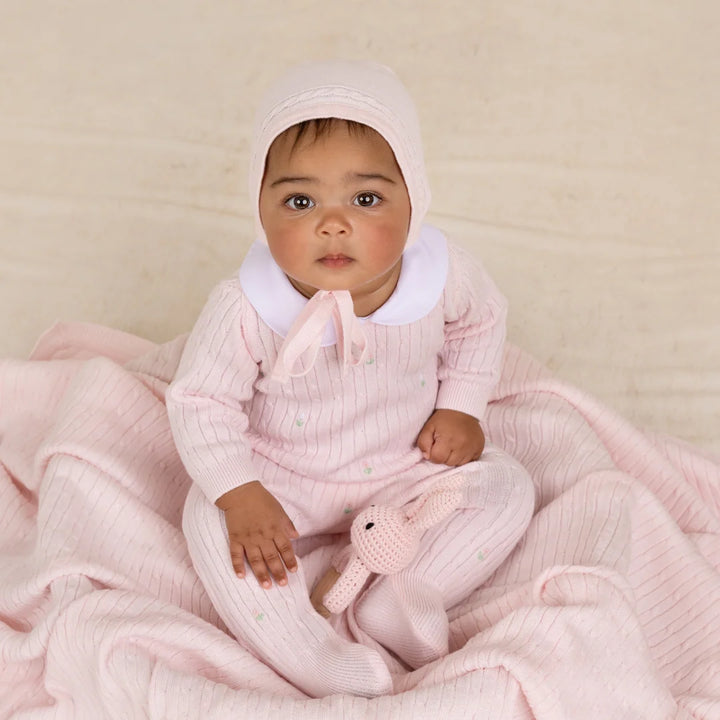 A baby wearing a light pink ribbed knit one-piece outfit with a white collar and matching bonnet, sitting on a soft pink blanket and holding a small pink stuffed bunny.