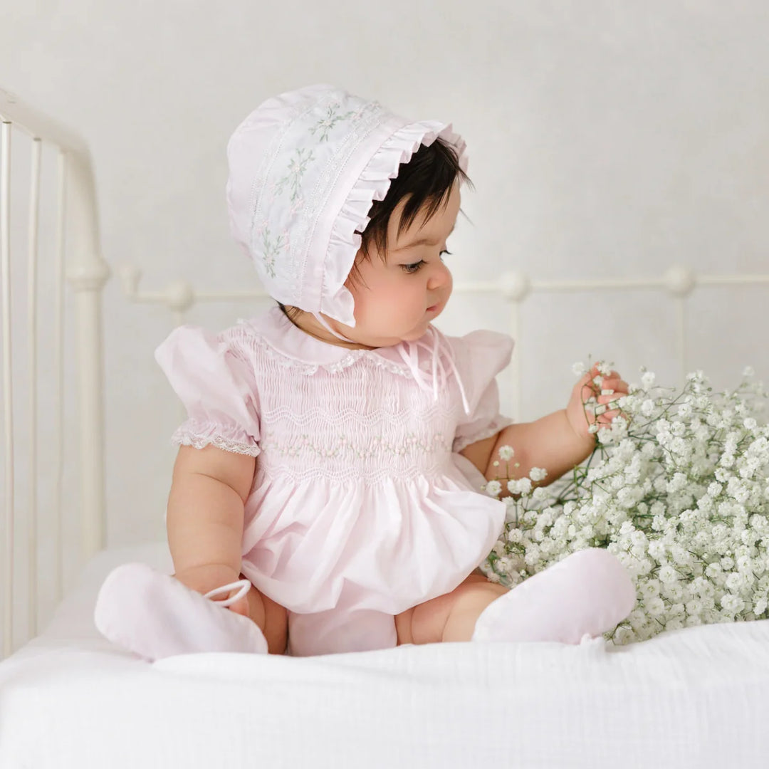 Baby in a pink outfit and white bonnet sitting on a bed with white flowers.