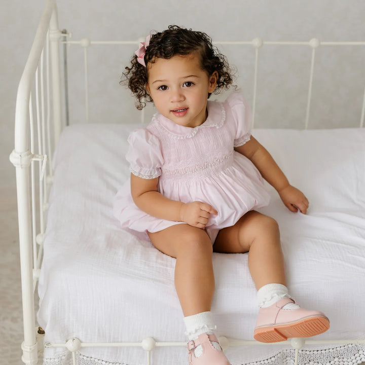 A toddler girl with curly brown hair sits on a white metal bed wearing a light pink smocked dress, white socks, and pink shoes.