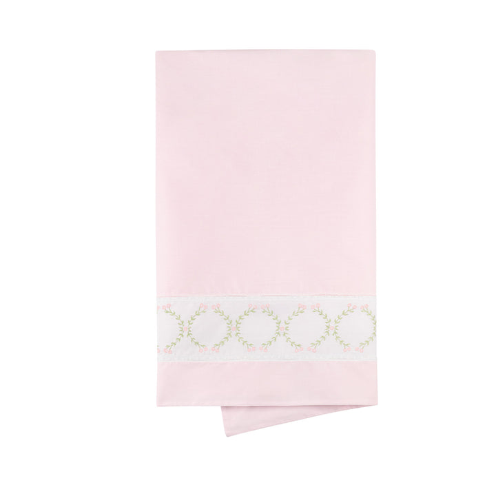 A folded light pink fabric item, likely a pillowcase or folded bed sheet, featuring a decorative white band near the bottom with a delicate pale green floral wreath pattern.