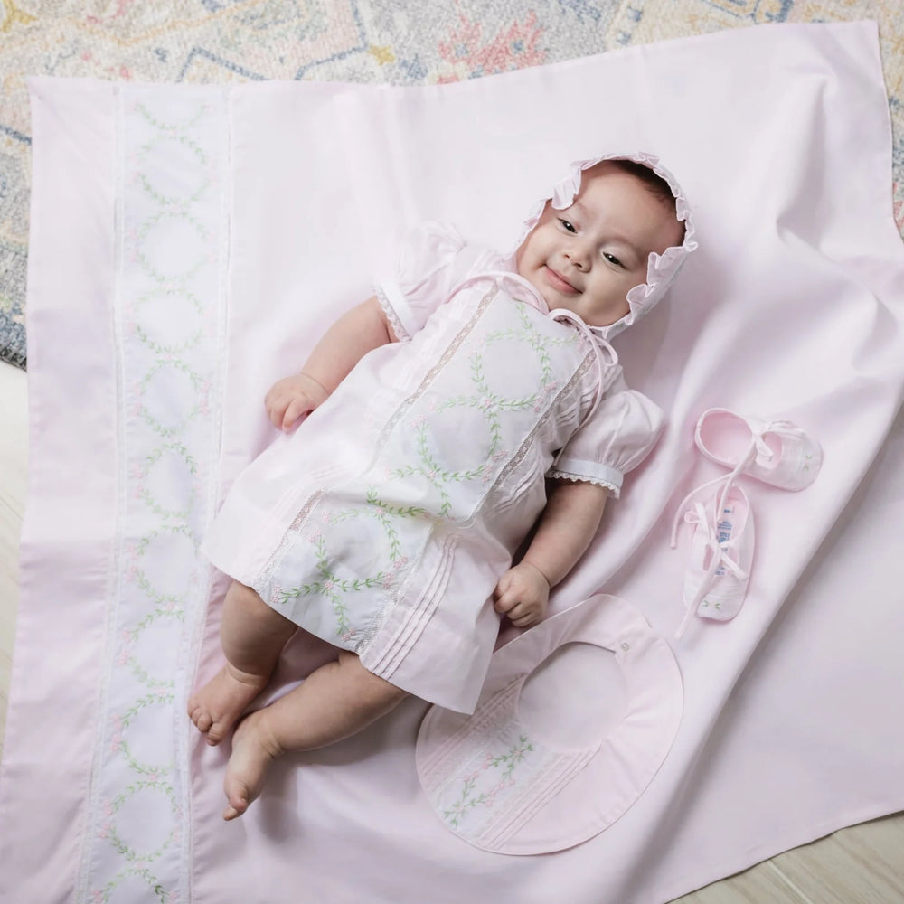 A baby wearing a light pink short-sleeve dress with white lace trim and green embroidered circular motifs, lying on a matching pink blanket. A matching pink bonnet and a small pink bootie are placed beside the baby.
