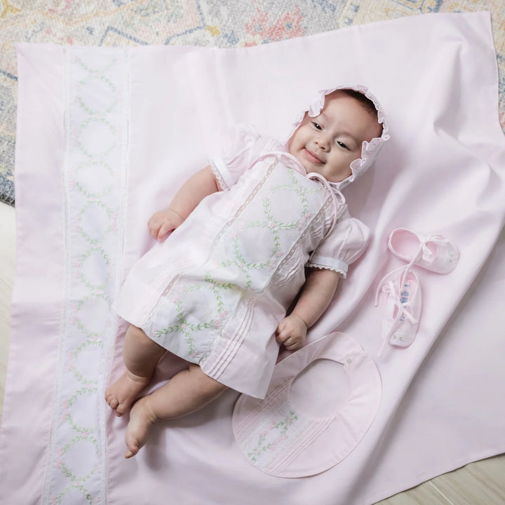 A baby wearing a light pink short-sleeve dress with white lace trim and green embroidered circular motifs, lying on a matching pink blanket. A matching pink bonnet and a small pink bootie are placed beside the baby.
