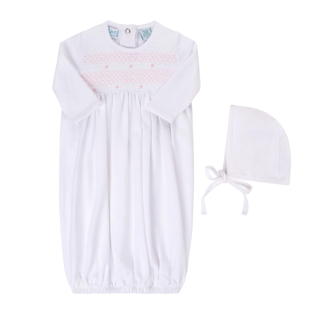A white baby smocked nightgown with long sleeves and a gathered smocked chest panel featuring light pink dotted lines and small pink hearts, shown with a matching white bonnet on a plain white background.