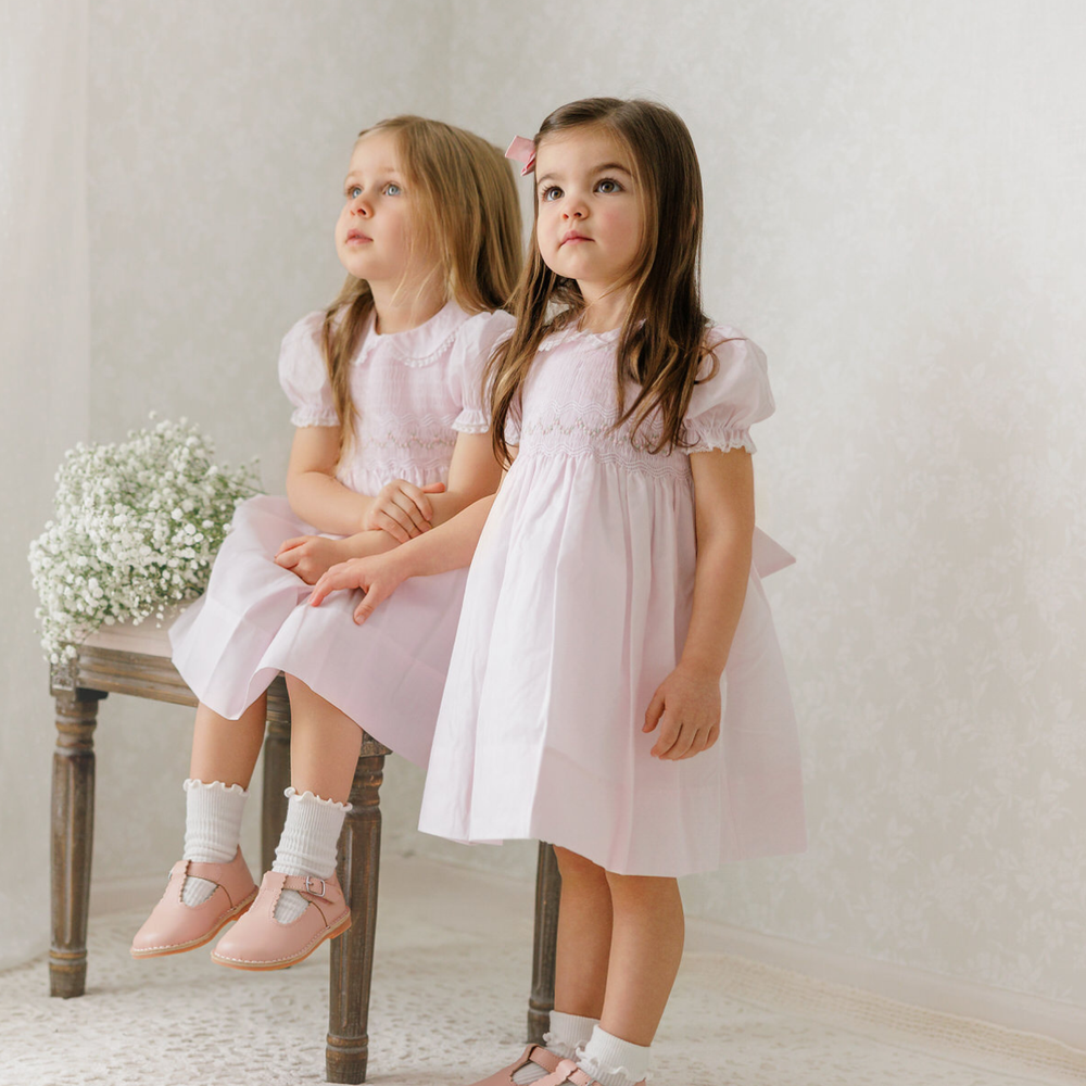 Two young girls in matching pink dresses sitting on a wooden stool with a white background.