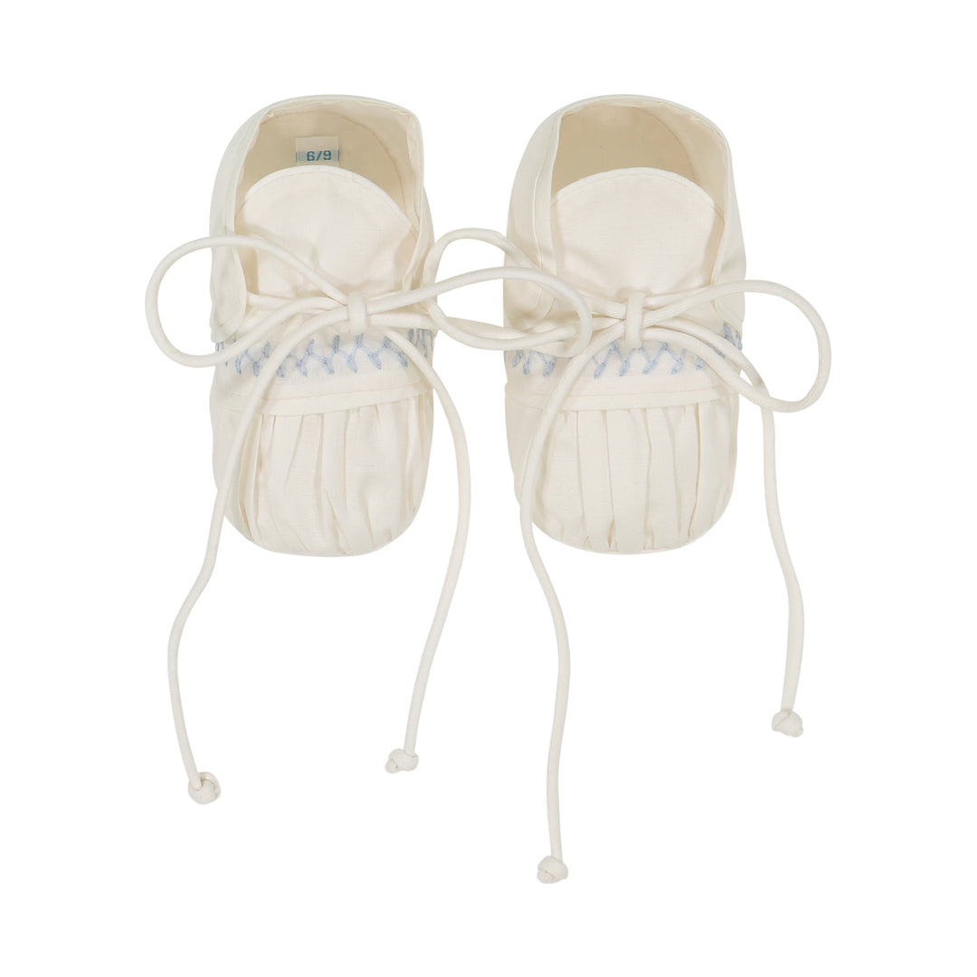 A pair of cream-colored baby moccasin shoes with white laces and a light blue patterned band near the top, shown against a plain white background.