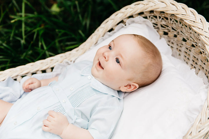 A baby wearing a light blue long-sleeve shirt and matching diaper cover lies on a white cushion inside a woven wicker bassinet outdoors, with green grass visible in the background.