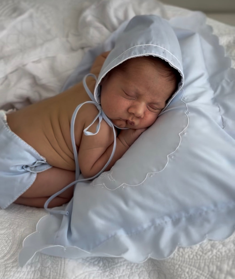 A sleeping newborn baby wearing a light blue bonnet and matching diaper cover, resting on a white pillow and white quilted bedding.