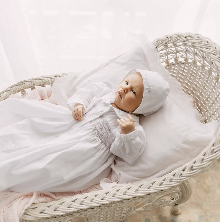 A baby wearing a white smocked christ gown and matching bonnet, lying on a white cushion inside a woven rattan bassinet.