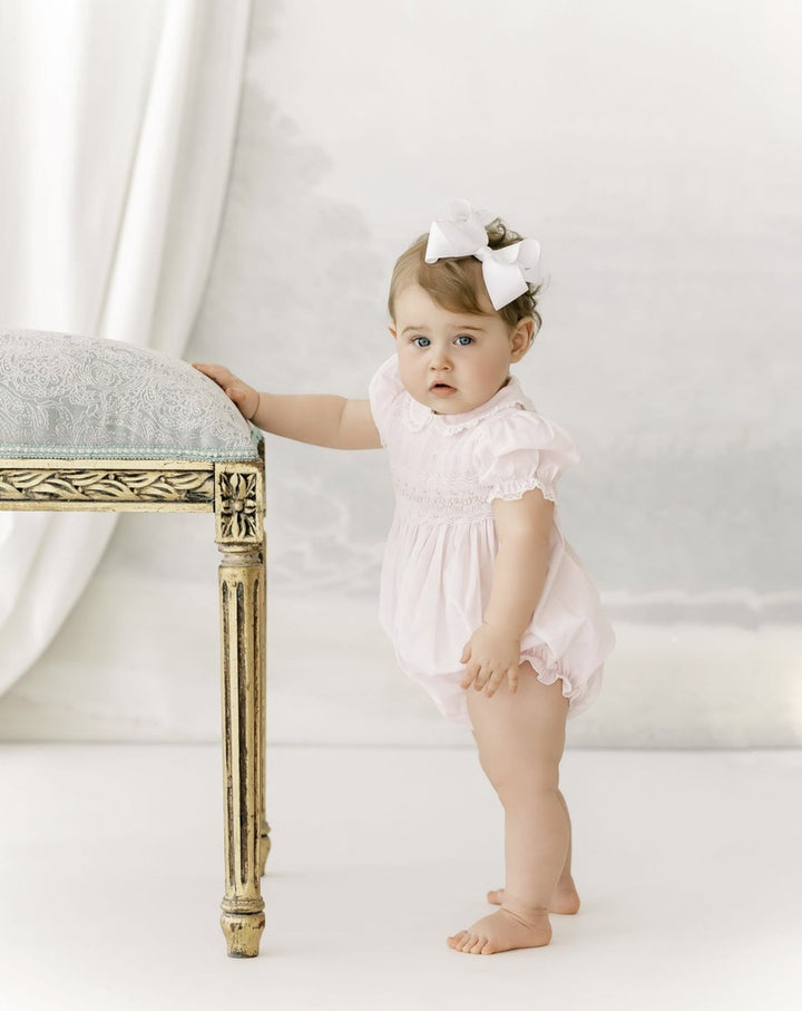 A baby girl wearing a light pink smocked romper and a white bow in her hair stands barefoot next to an ornate gold bench in a soft, high-key studio setting.
