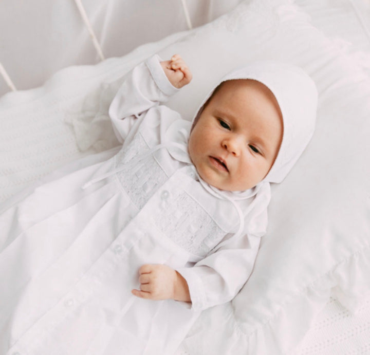A baby lying on white bedding, wearing a white long-sleeve baby outfit with a smocked chest panel and a matching bonnet.