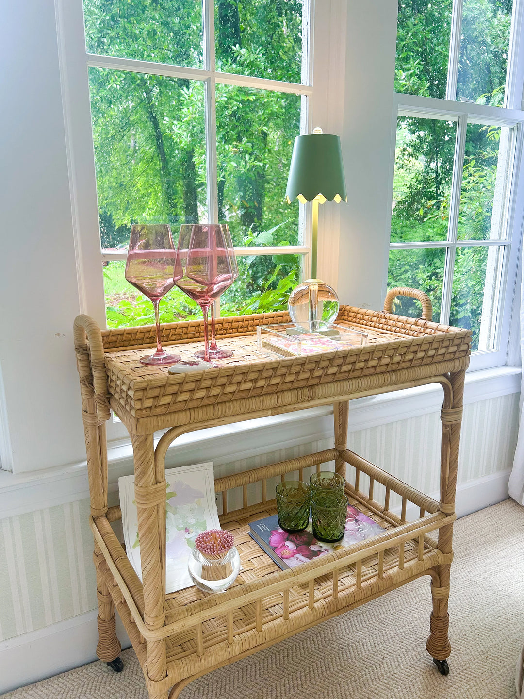 Wicker bar cart with wine glasses and decor items in a room with large windows.