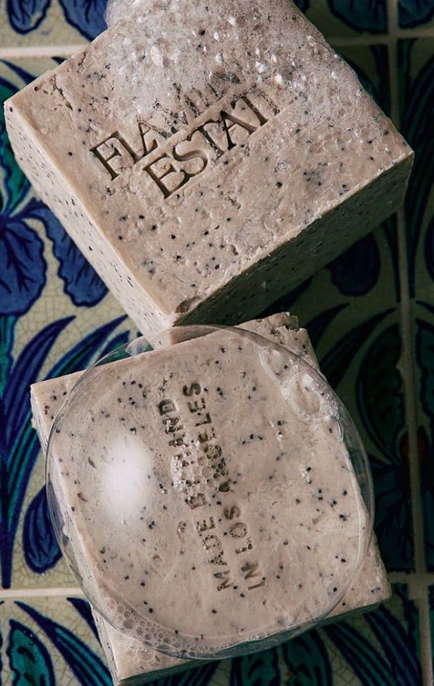 Two rectangular bars of speckled soap resting on a blue and cream patterned tile surface. One bar is upright and embossed with the brand name 'FLORIDA ESTATI', while the other lies flat with a clear plastic cap covering part of it.