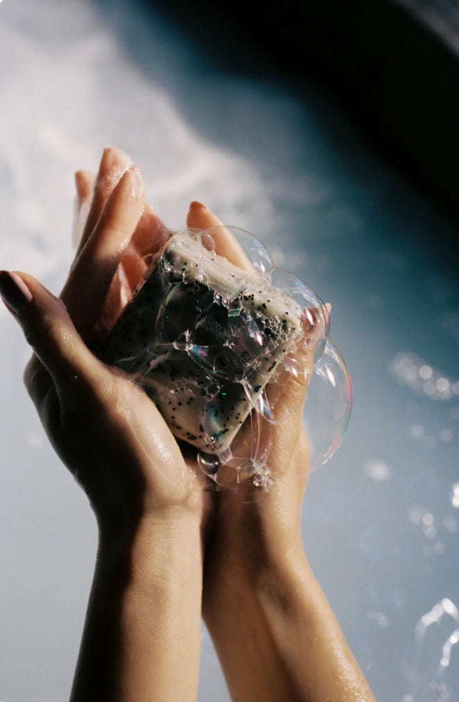 A close-up of hands holding a decorative soap bar wrapped in clear bubbles, set against a dark, moody background with water droplets.