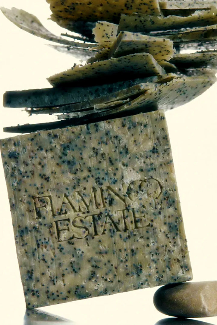 A close-up of a speckled stone-textured bar of soap standing upright, with layered flat stones stacked on top and a smooth round stone at the base. The soap is embossed with the words 'FLAMINGO ESTATE'.