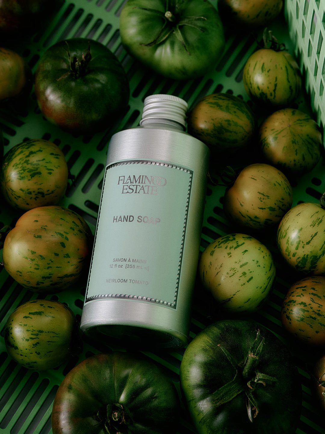 Hand soap bottle labeled 'Flamingo Estate' surrounded by green tomatoes on a green surface.