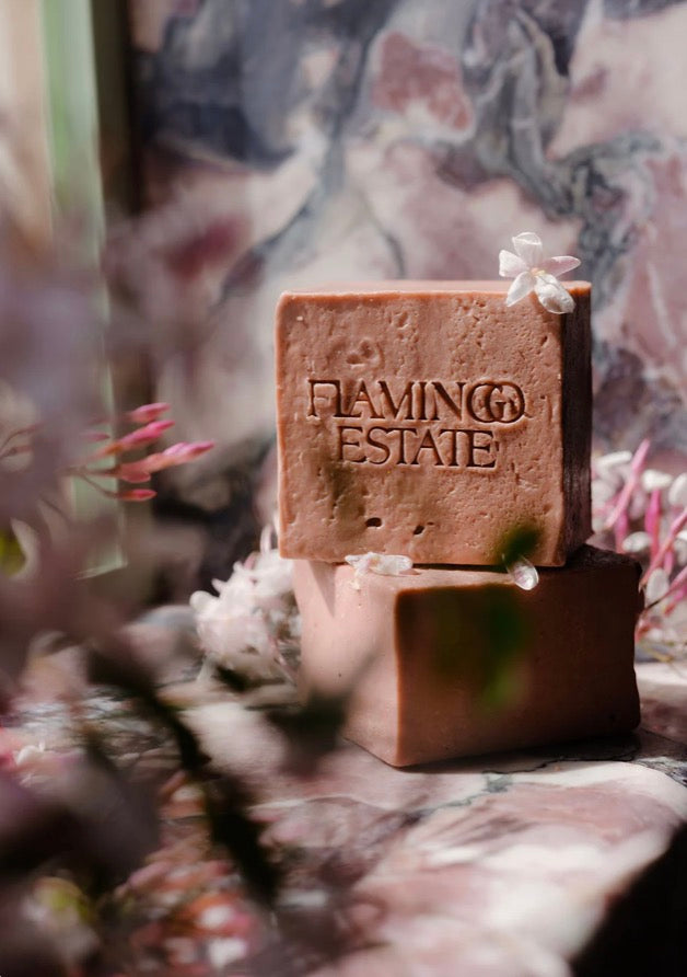 Two stacked rectangular bars of soap in a warm brownish-terracotta color, with the top bar embossed with the brand name 'FLAMING ESTATE'. The bars are styled on a marbled surface with soft, out-of-focus botanical elements in the foreground.
