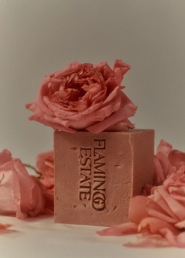 A rectangular bar of soap with the brand name 'FLAMING ESTATE' embossed vertically, topped with a large pink rose, surrounded by scattered pink rose petals on a neutral light gray background.
