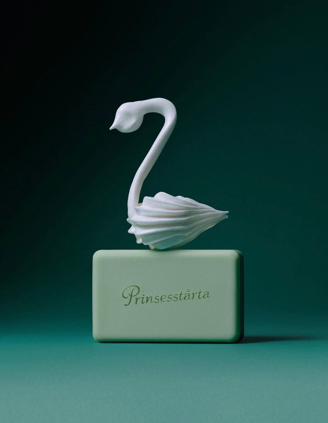 A white swan figurine perched on a rectangular pale green base engraved with the brand name 'Prinesstärta', set against a dark teal studio background.