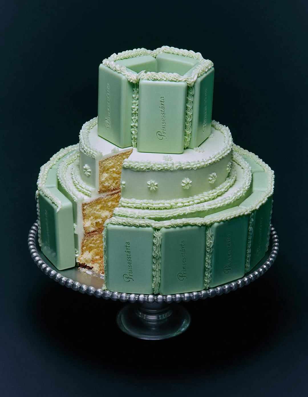 A decorative tiered cake designed to look like a chocolate bar, with pale green and white frosting, set on a silver pedestal against a dark background. A slice is cut from the side, showing a golden cake layer.