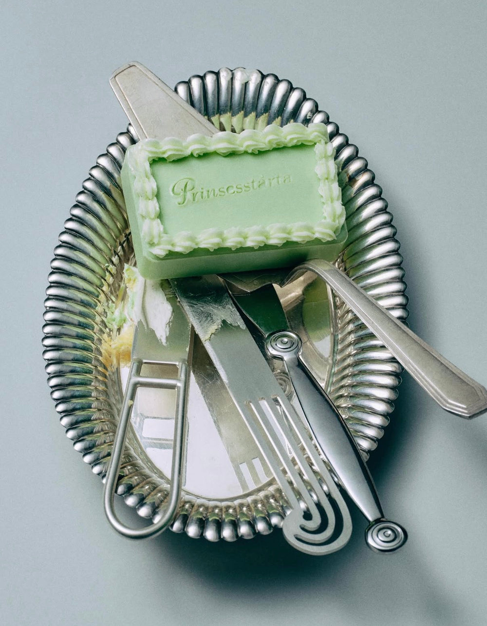A light green Prinsesstone soap bar with embossed lettering, arranged on top of a silver scalloped dish with cutlery, against a neutral gray background.