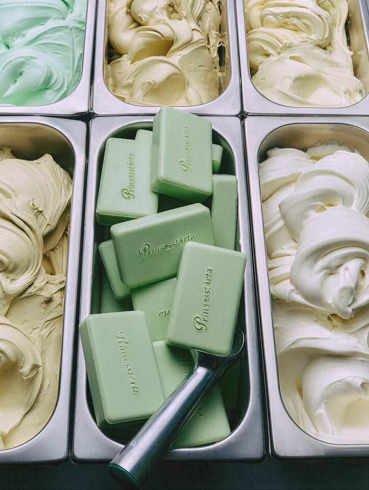 A top-down view of an ice cream or gelato shop display with multiple metal bins filled with creamy swirls in mint green, pale yellow, and white. In the center bin, pale green branded bars are stacked with a metal scoop resting on top.