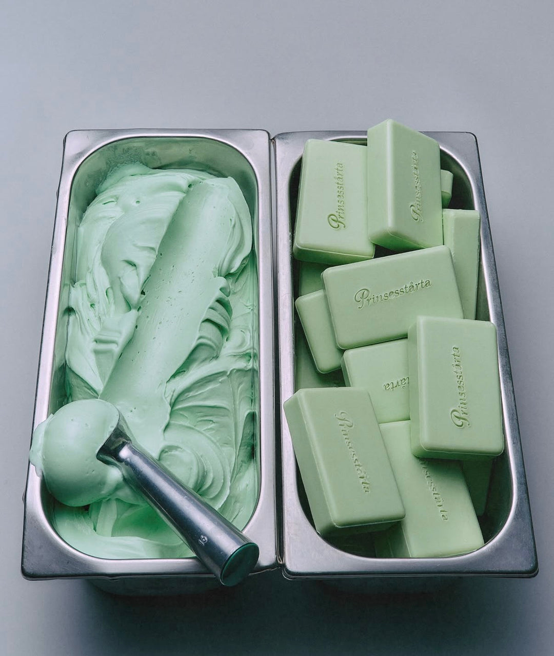 A top-down view of a mint green ice cream tub with a metal scoop next to a metal container filled with matching mint green bars of soap, all on a neutral gray background.