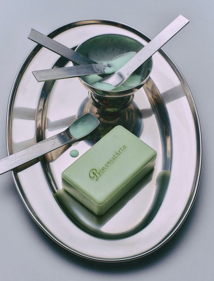 A rectangular pale green soap bar with the brand name 'Pierrefitte' embossed on top, displayed on a large oval silver metal tray alongside matching silver tools and a small bowl.
