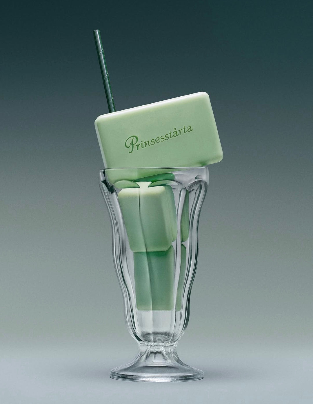 A pale mint green rectangular bar of soap with the brand name 'Prunssestárta' embossed on it, resting inside a clear fluted glass with a dark green straw, set against a dark gray gradient background.