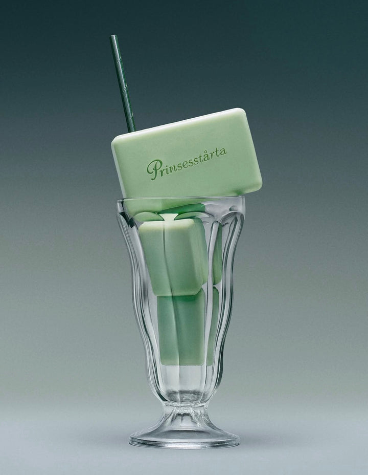 A pale mint green rectangular bar of soap with the brand name 'Prunssestárta' embossed on it, resting inside a clear fluted glass with a dark green straw, set against a dark gray gradient background.