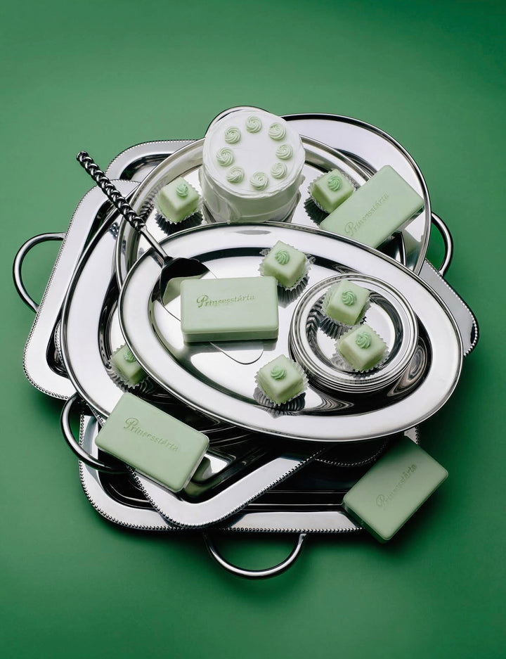 A styled product shot of a collection of pale mint green soaps and bath products arranged on a stack of overlapping silver metal trays against a solid green background. The brand name 'Bourgeon' is embossed on several bars.