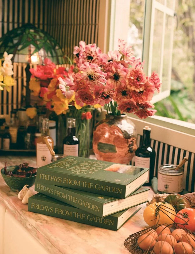 Two stacked books titled 'FRIDAYS FROM THE GARDEN' sit on a windowsill surrounded by pink flowers, small bottles, eggs, and pumpkins in a warm, autumnal setting.