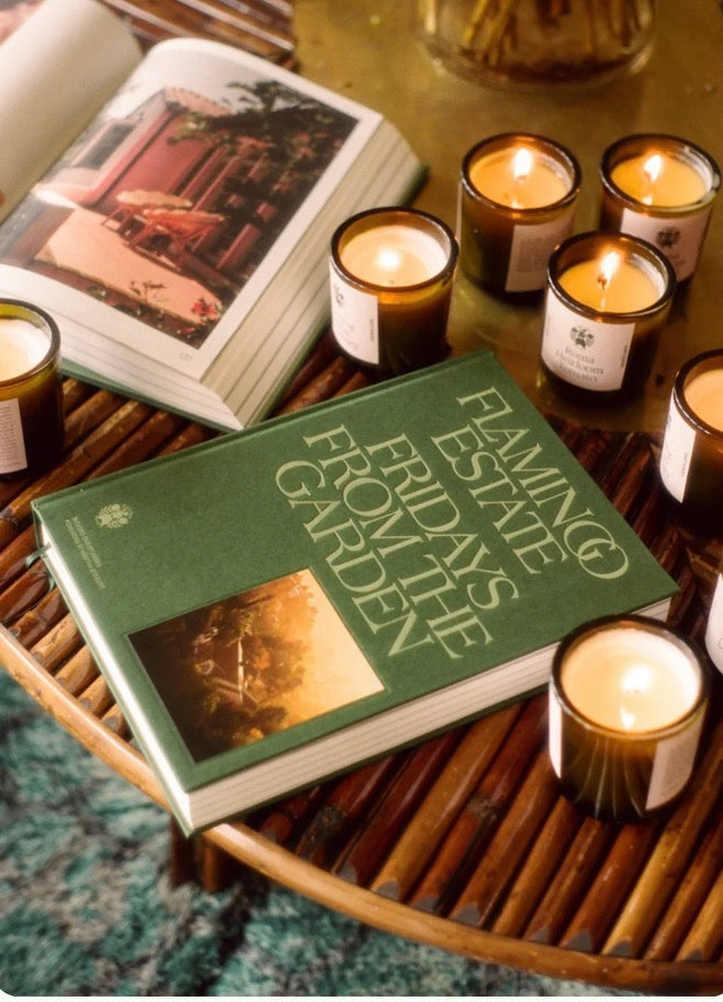 A green hardcover book titled 'Flamingo Estate Fridays from the Garden' styled on a round woven tray with lit candles and an open magazine nearby.